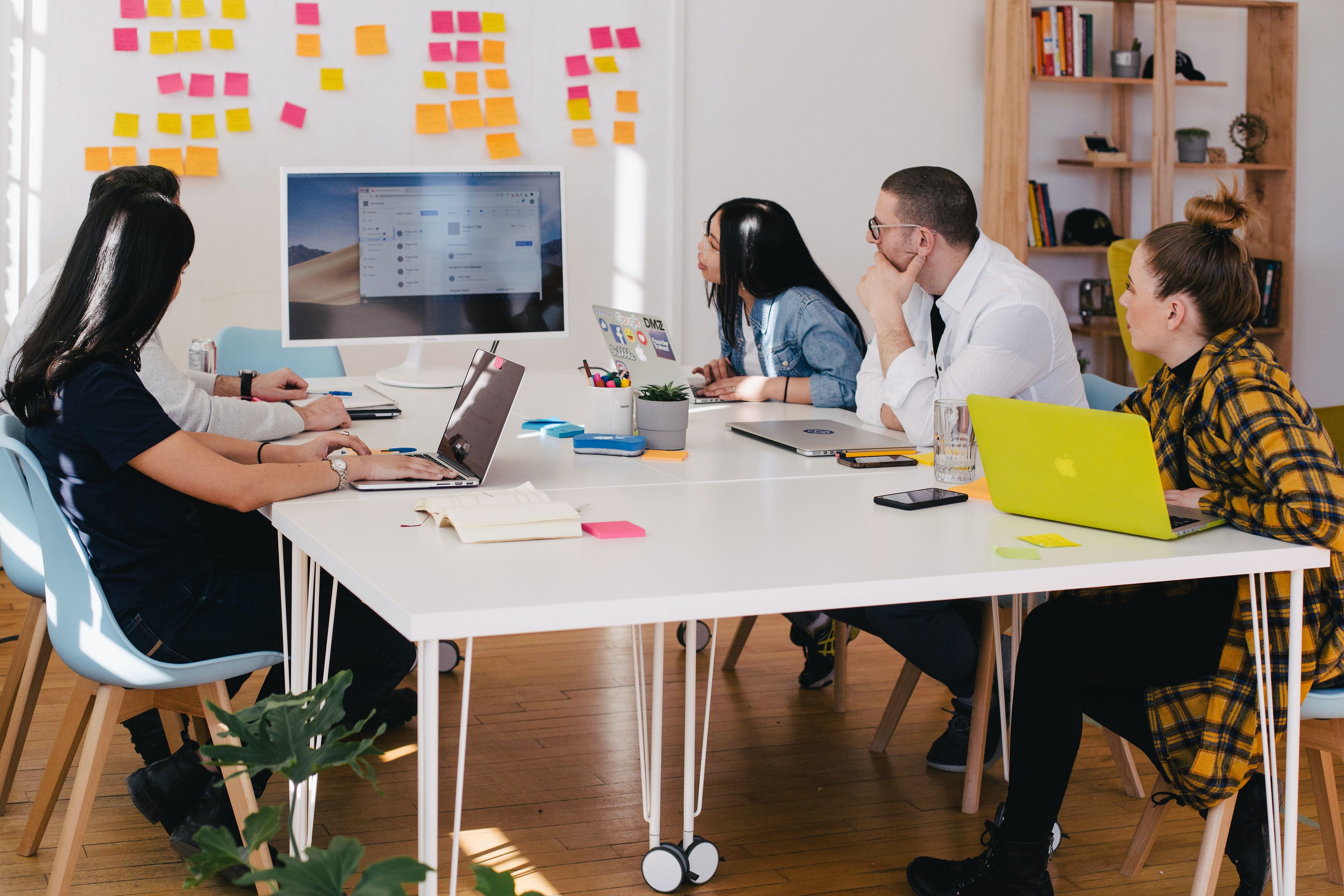 Five people in a meeting around a table all looking toward the screen that is on the table. Behind that screen are post-its on the wall.