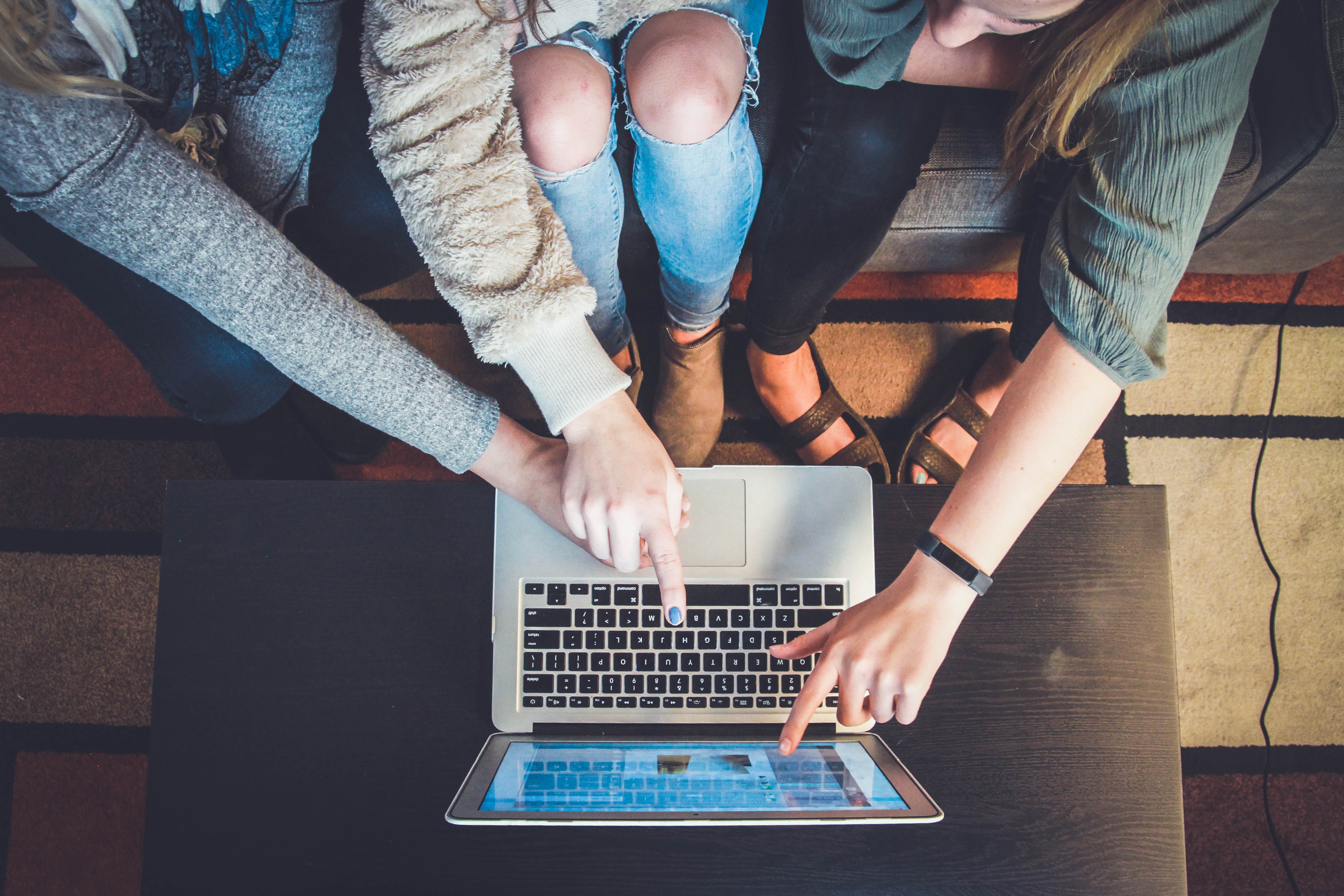 Top view of four people sitting around a laptop, three of them pointing to the laptop's screen.