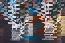 Top view of four people sitting around a laptop, three of them pointing to the laptop's screen.