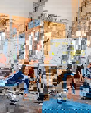 A presenter stands in front of a whiteboard on which a table has been drawn and post-its have been added into some cells. In the picture, three others are listening intently.